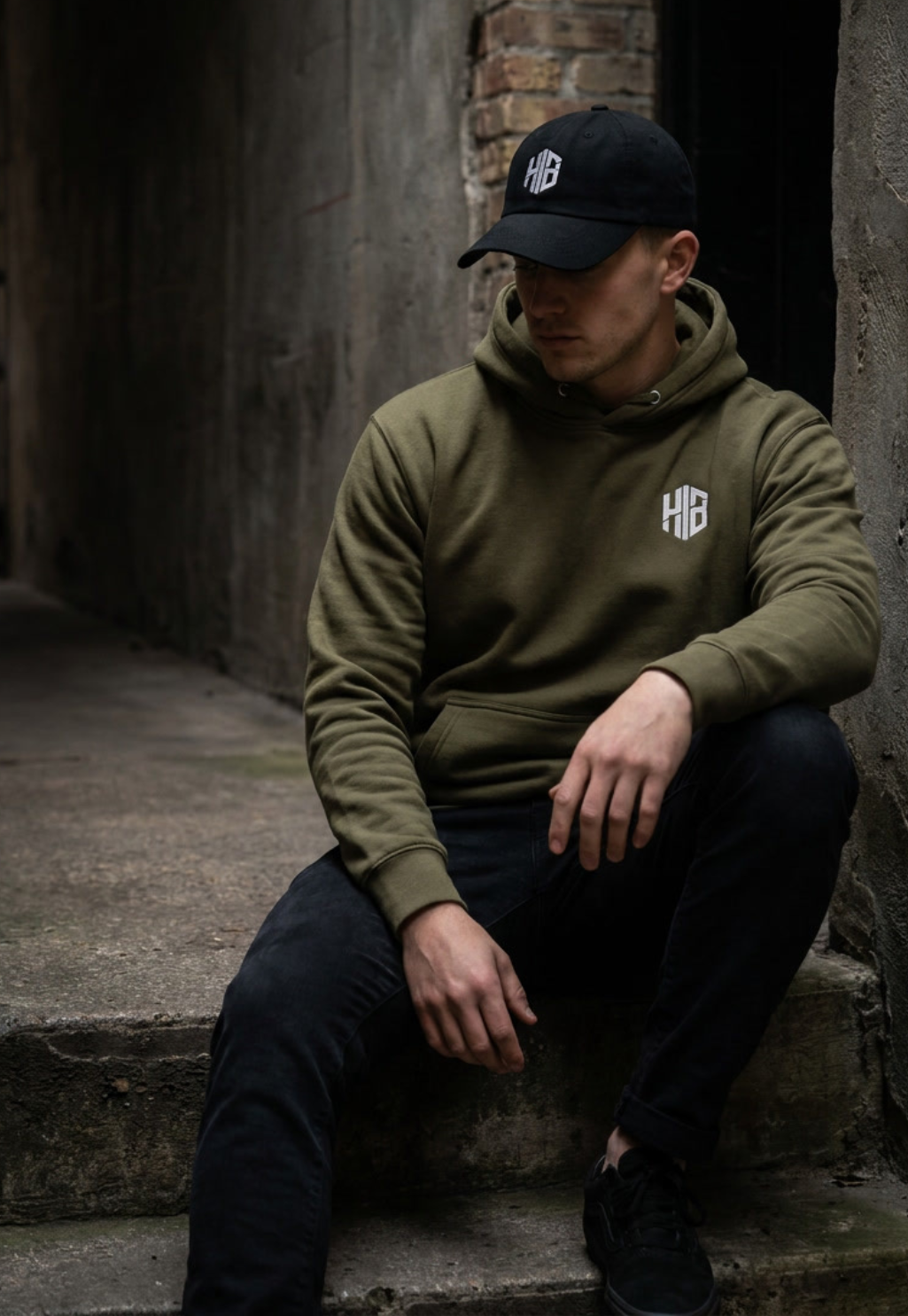 Male model wearing a HELVEN BOR embroidered hoodie and black cap in an urban streetwear setting